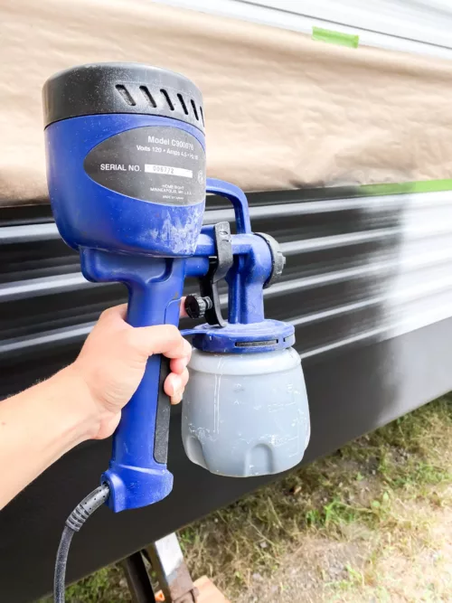 A hand holding a paint sprayer with an RV exterior in the background